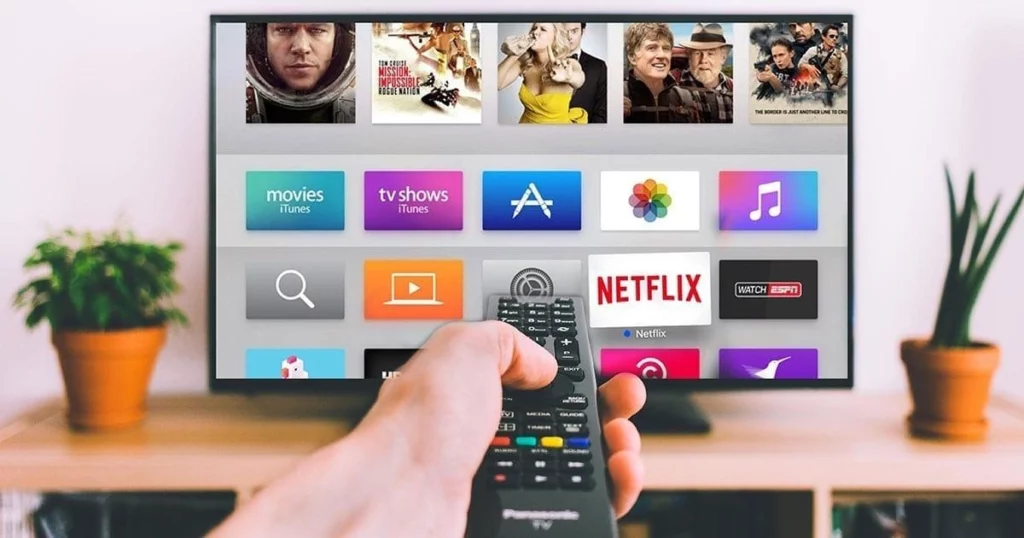 Hand holding remote pointed at TV screen displaying streaming apps like Netflix, iTunes movies, and ESPN in a cozy living room.